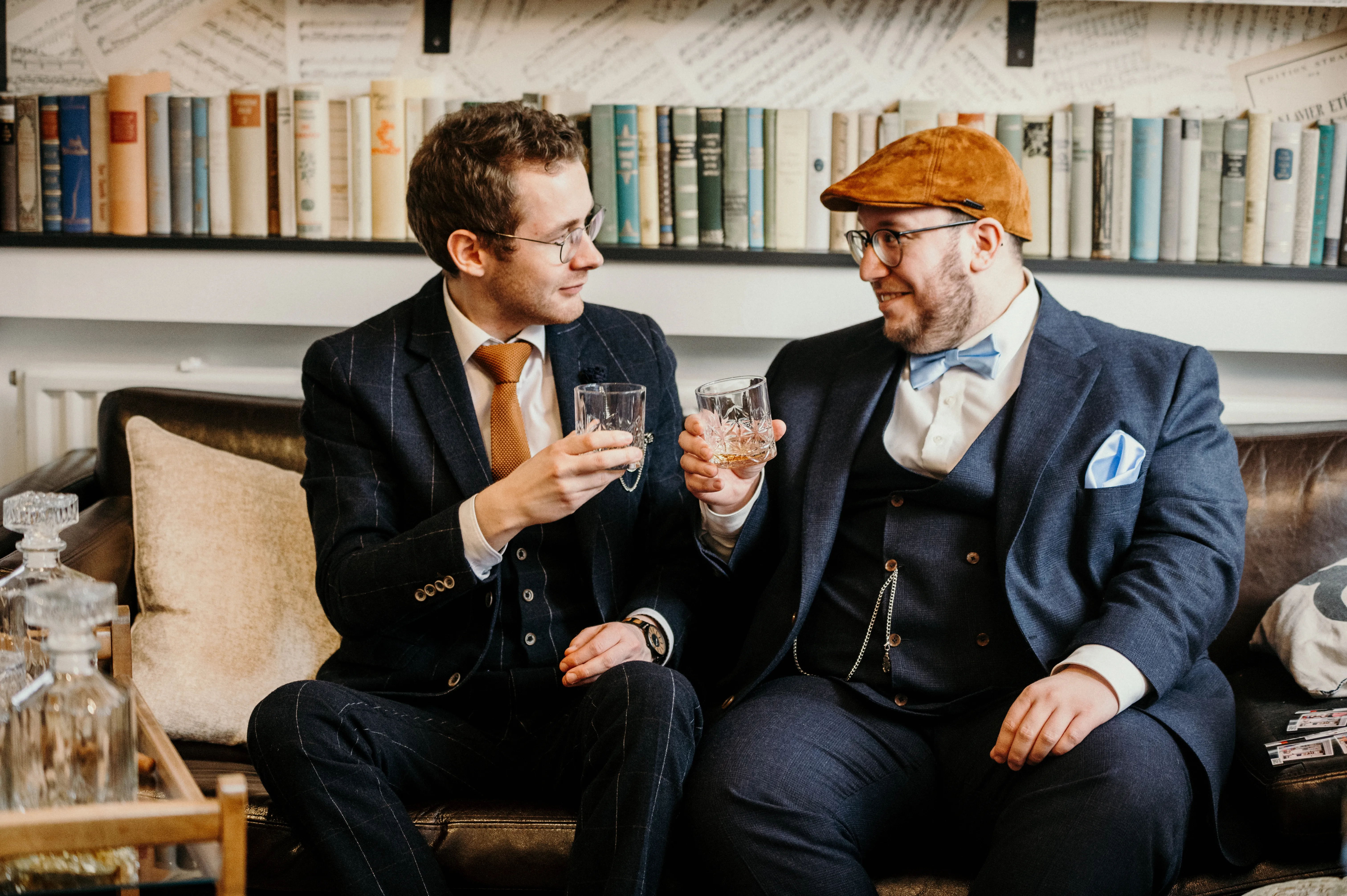 Zwei Männer in Anzügen stoßen mit Whiskygläsern auf einem Sofa vor einem Bücherregal an