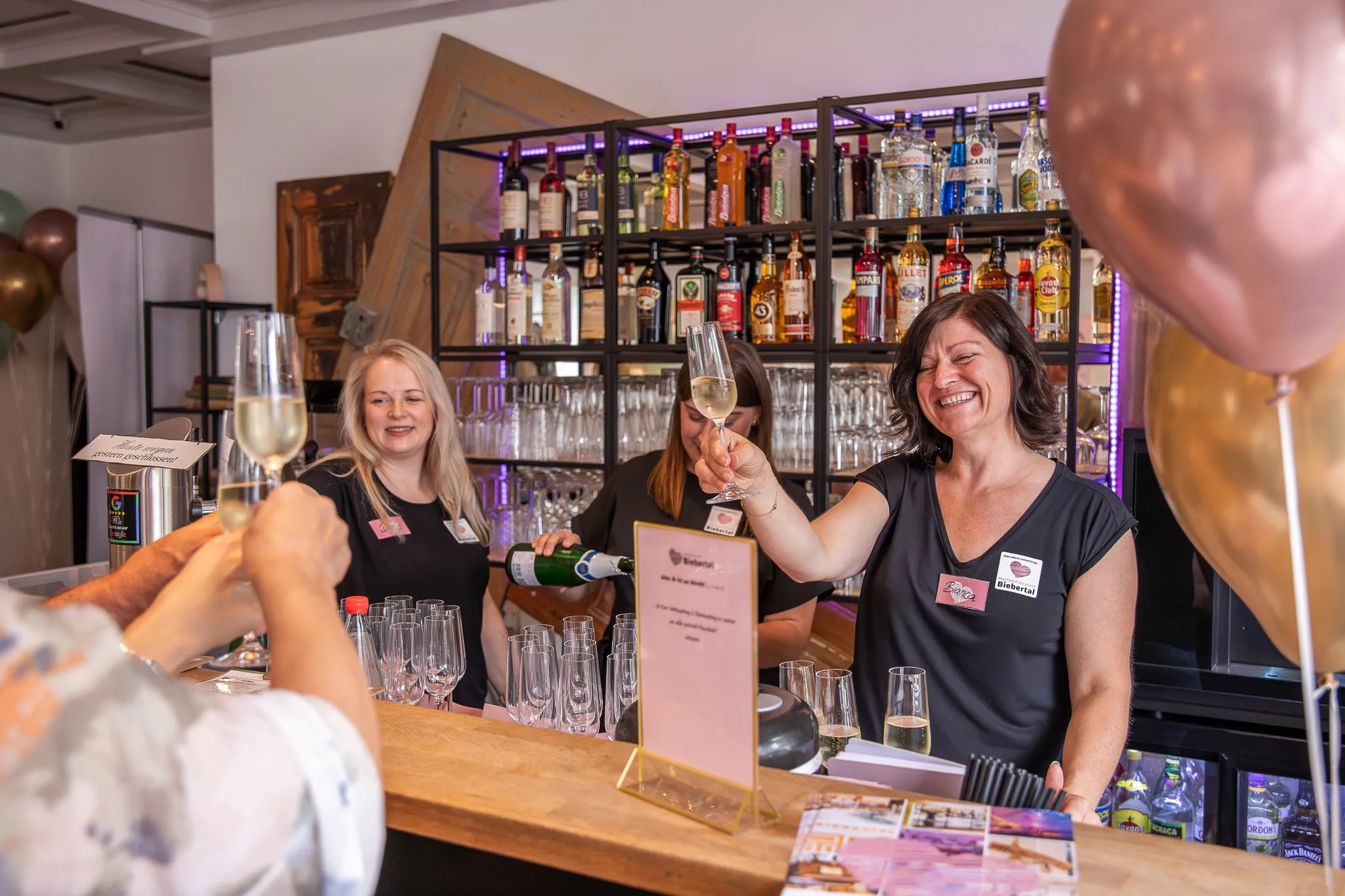 Barteam stößt mit Sektgläsern an der Bar mit beleuchtetem Spirituosenregal an