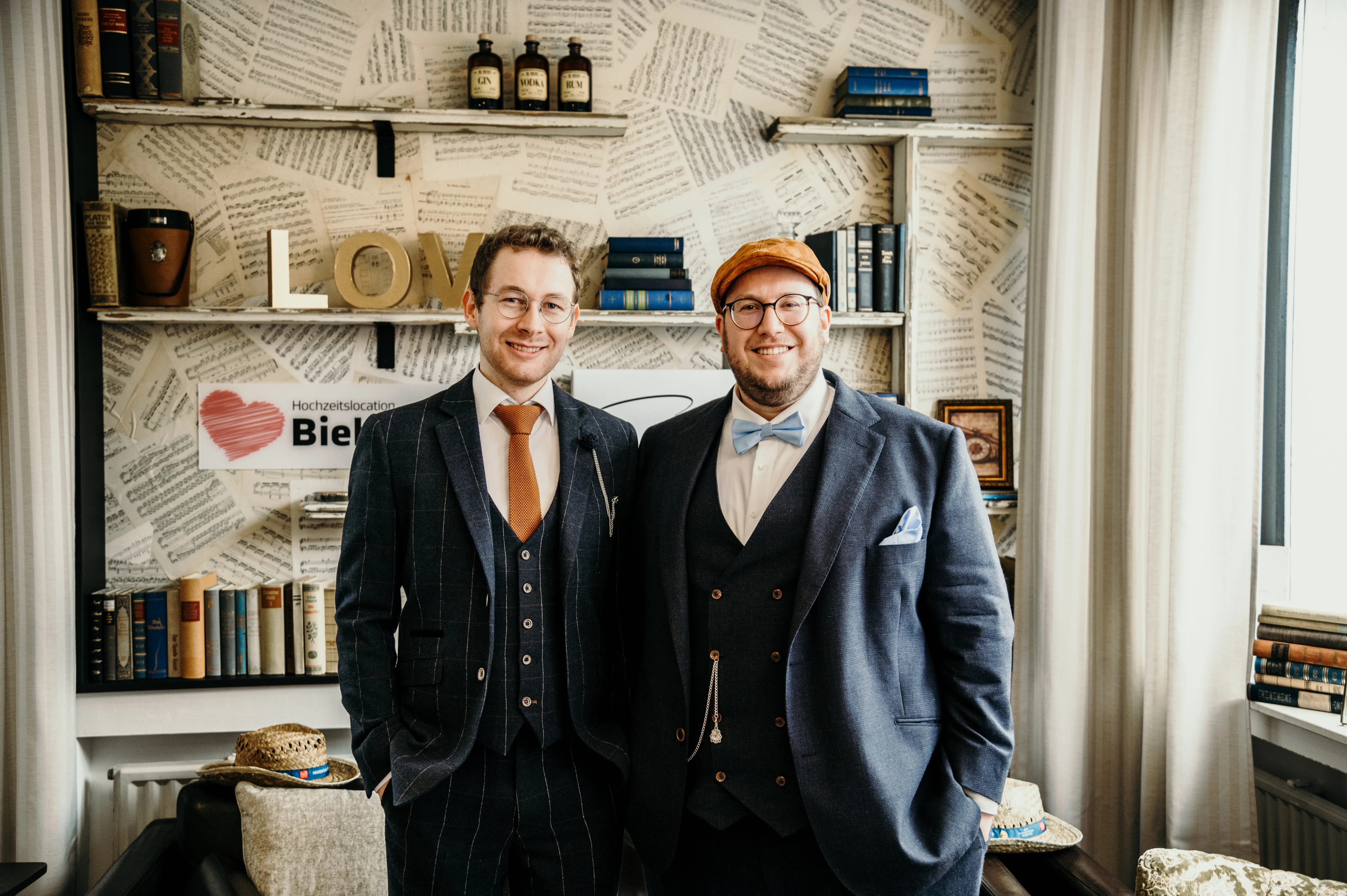 Zwei Männer in Vintage-Anzügen vor Notenblatt-Wand mit Bücherregal in der Hochzeitslocation Biebertal