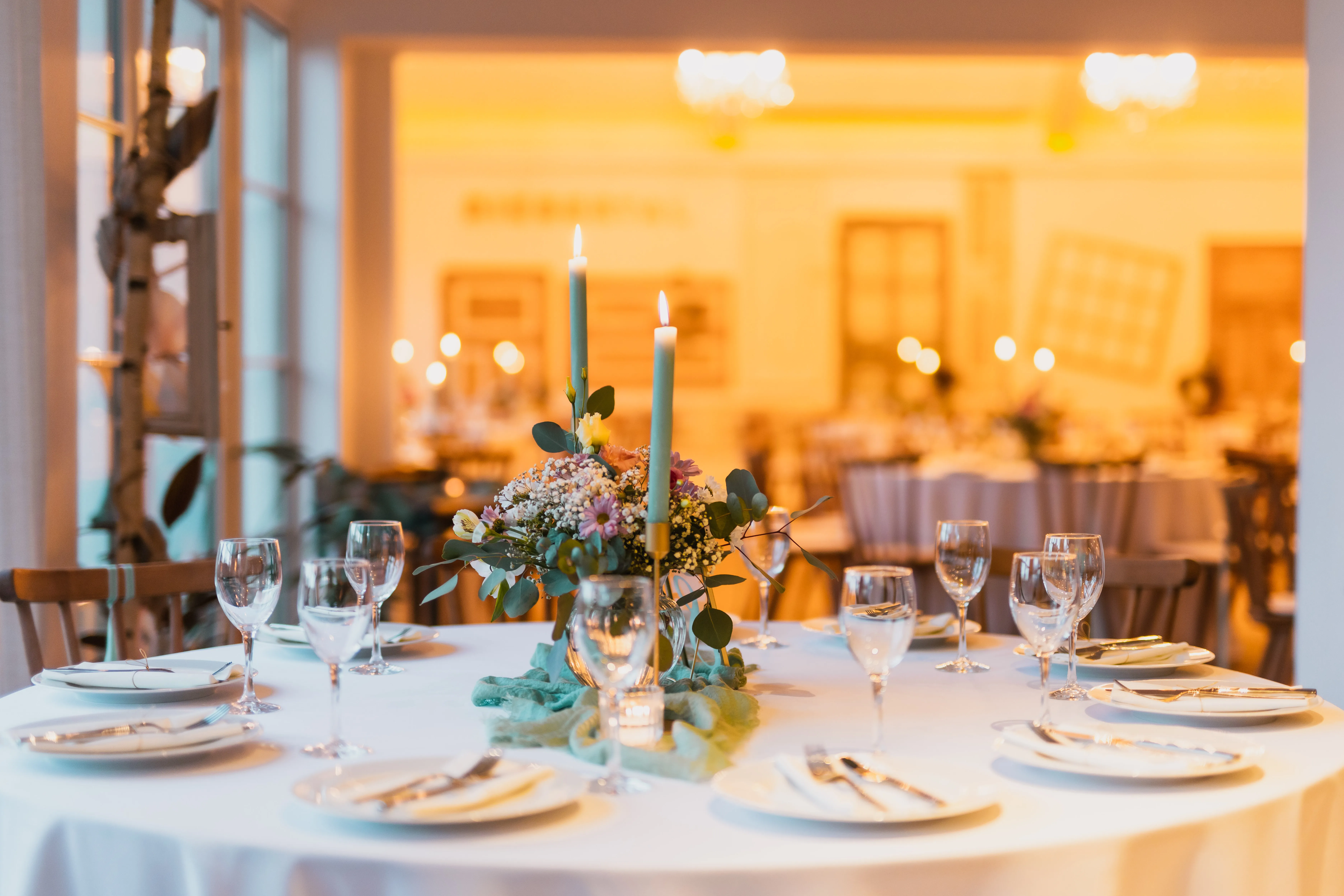 Eingedeckter runder Hochzeitstisch mit Blumen und Kerzen, dahinter der beleuchtete Festsaal