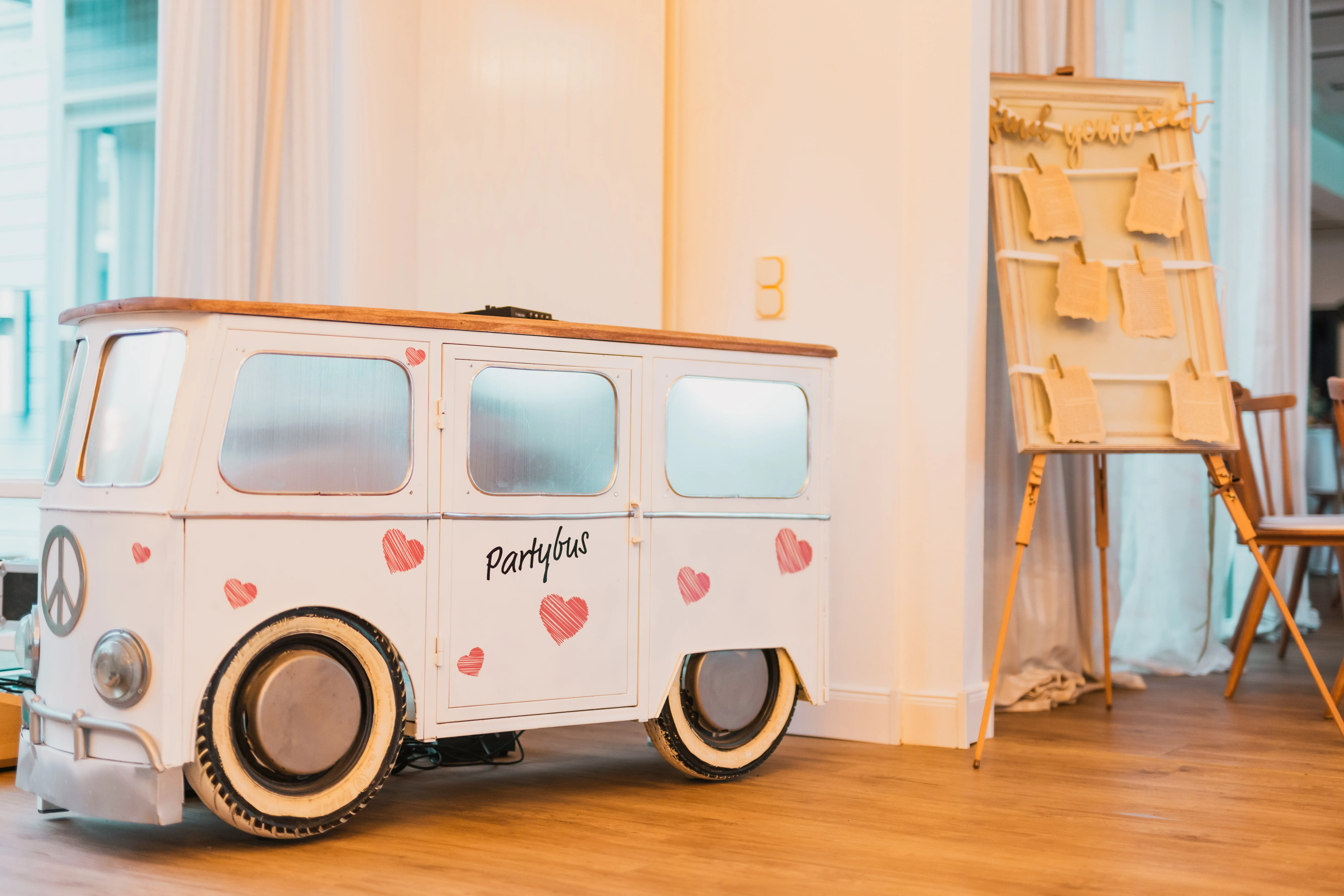 Weißer Vintage-Partybus im Bulli-Stil mit roten Herzen als Dekoelement, daneben Sitzplan-Staffelei