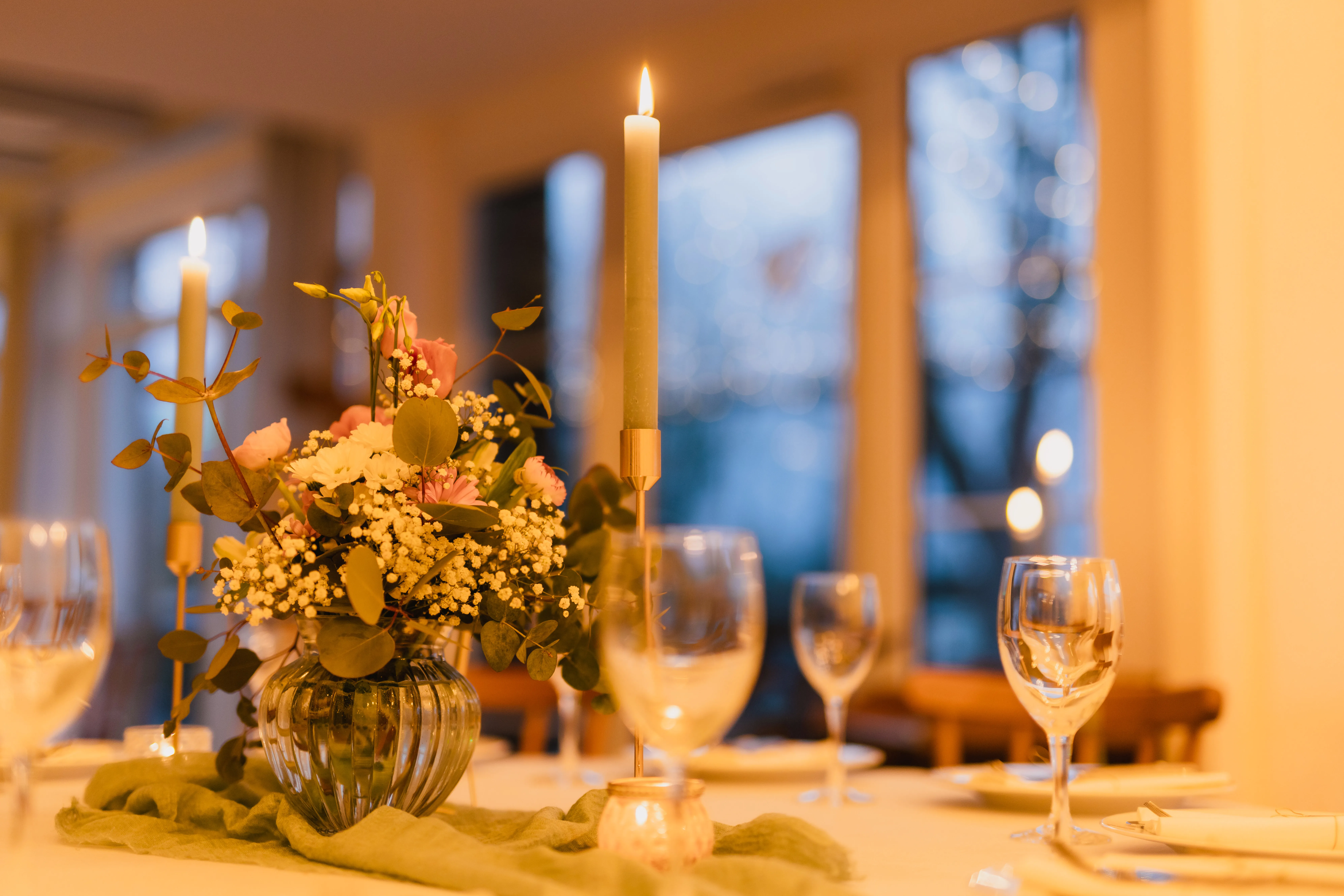 Tischdekoration mit Blumengesteck, brennenden Kerzen und Weingläsern zur blauen Stunde im Festsaal